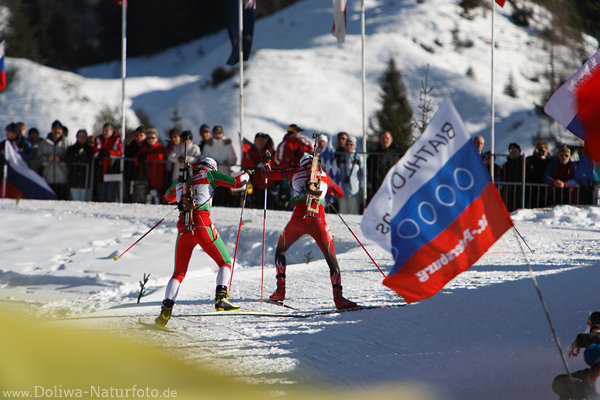 iathleten Paar im Skilauf zwischen Fahnen der Zuschauer an Loipe im Biathlonstadion