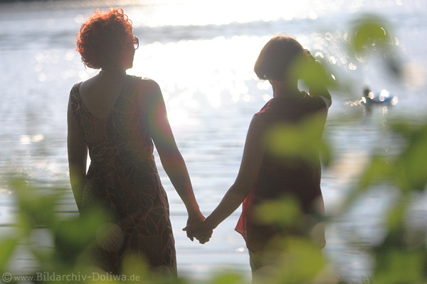 Jenseitslicht Sonnenreflexe über Frauenpaar Silhouetten Hand in Hand am Wasser