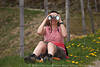 1201628_Mädchen lustiges Bild durch Fernglas schauen lachen Naturporträt Bergwandererin im Gras sitzen am Zaun