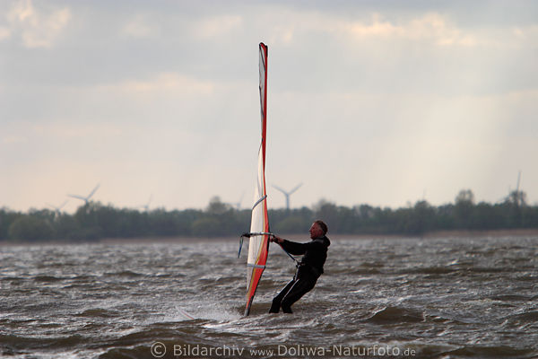 Windsurfer an Windsegel tief in Wasser stehend gegen Wind auf See herausfahren seitlich am Rigg